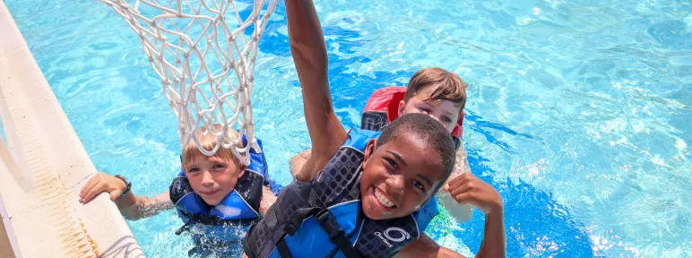 Three boys in lifejackets playing basketball in the pool
