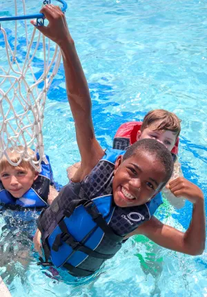 Three boys in lifejackets playing basketball in the pool